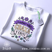 Футболка найкраща сваха, білого кольору, складена на столі, фон світло-сірий, принт з написом 'НАЙКРАЩА СВАХА У СВІТІ' в пастельних кольорах, з біркою на зеленій стрічці.