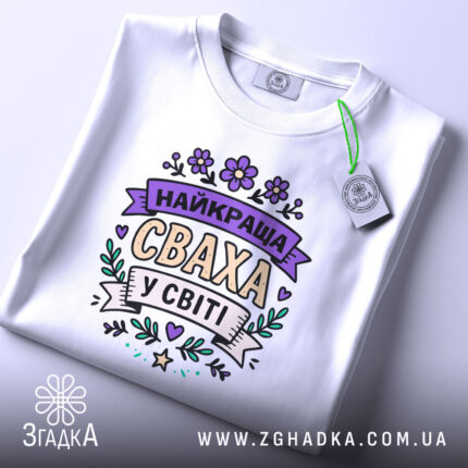 Футболка найкраща сваха, білого кольору, складена на столі, фон світло-сірий, принт з написом 'НАЙКРАЩА СВАХА У СВІТІ' в пастельних кольорах, з біркою на зеленій стрічці.