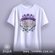 Футболка найкраща сваха, білого кольору, підвішена на вішалці, фон світло-сірий, принт з написом 'НАЙКРАЩА СВАХА У СВІТІ' в пастельних кольорах з квітковими елементами.