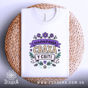 Футболка найкраща сваха, білого кольору, складена на кошику, фон світло-сірий, принт з написом 'НАЙКРАЩА СВАХА У СВІТІ' в пастельних кольорах, на плетеній основі.