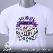 Футболка найкраща сваха, білого кольору, складена на столі, фон світло-сірий, принт з написом 'НАЙКРАЩА СВАХА У СВІТІ' в пастельних кольорах, з біркою на зеленій стрічці.