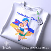 Футболка найкраща сваха, біла, складена на столі, фон світлий, принт з дівчиною в українському вбранні, яка тримає качалку і кошик з солодощами, на фоні контур України з жовто-синім кольорами, з етикеткою.