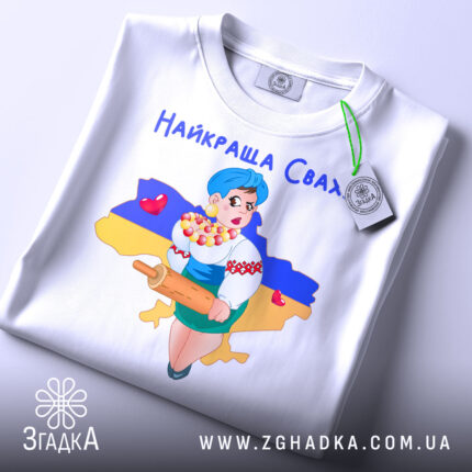 Футболка найкраща сваха, біла, складена на столі, фон світлий, принт з дівчиною в українському вбранні, яка тримає качалку і кошик з солодощами, на фоні контур України з жовто-синім кольорами, з етикеткою.