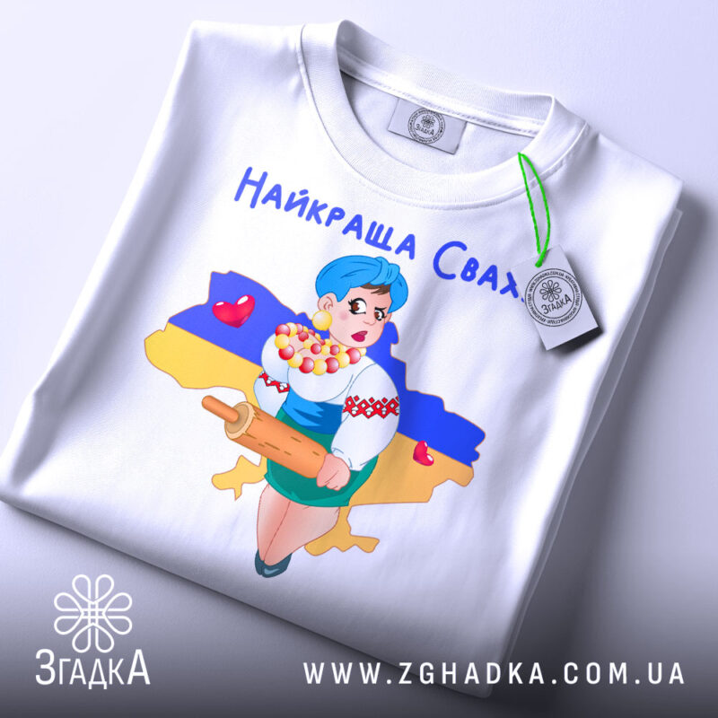 Футболка найкраща сваха, біла, складена на столі, фон світлий, принт з дівчиною в українському вбранні, яка тримає качалку і кошик з солодощами, на фоні контур України з жовто-синім кольорами, з етикеткою.