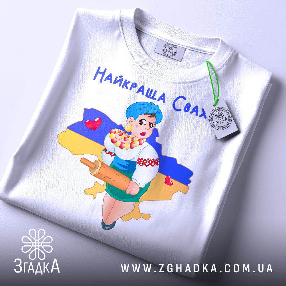 Футболка найкраща сваха, біла, складена на столі, фон світлий, принт з дівчиною в українському вбранні, яка тримає качалку і кошик з солодощами, на фоні контур України з жовто-синім кольорами, з етикеткою.