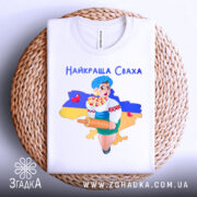 Футболка найкраща сваха, біла, складена на плетеній основі, фон нейтральний, принт з дівчиною в українському вбранні, яка тримає качалку і кошик з солодощами, на фоні контур України з жовто-синім кольорами.