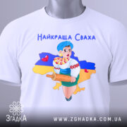 Футболка найкраща сваха, біла, складена на столі, фон світлий, принт з дівчиною в українському вбранні, яка тримає качалку і кошик з солодощами, на фоні контур України з жовто-синім кольорами, з етикеткою.