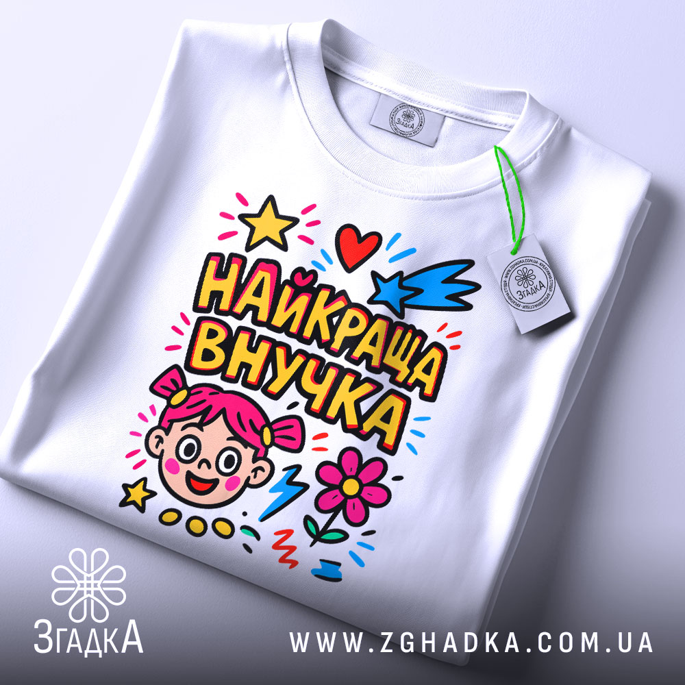 Футболка найкраща внучка, біла, зображена зверху, складена на поверхні, фон світло-сірий, принт кольоровий з малюнком дівчинки, зірочками, серцями та квітами, з етикеткою на комірці.