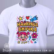 Футболка найкраща внучка, біла, зображена зверху, складена на поверхні, фон світло-сірий, принт кольоровий з малюнком дівчинки, зірочками, серцями та квітами, з етикеткою на комірці.