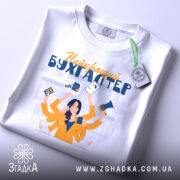 Футболка найкращий бухгалтер, біла, складена на поверхні, фон світлий, принт з жінкою в жовтій блузці, яка тримає документи та гаджети, з етикеткою на комірці.