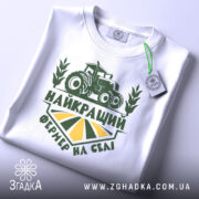 Футболка найкращий фермер, біла, складена на поверхні, фон світлий, принт зелений з трактором та написом 'НАЙКРАЩИЙ ФЕРМЕР НА СЕЛІ', з етикеткою на рукаві.