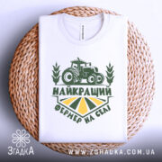 Футболка найкращий фермер, біла, складена на плетеній основі, фон світлий, принт зелений з трактором та написом 'НАЙКРАЩИЙ ФЕРМЕР НА СЕЛІ'.