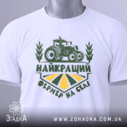 Футболка найкращий фермер, біла, складена на поверхні, фон світлий, принт зелений з трактором та написом 'НАЙКРАЩИЙ ФЕРМЕР НА СЕЛІ', з етикеткою на рукаві.