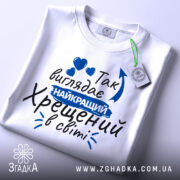 Футболка найкращий хрещений, біла, зображена зверху, фон світло-сірий, принт з текстом синього кольору, на футболці є етикетка.