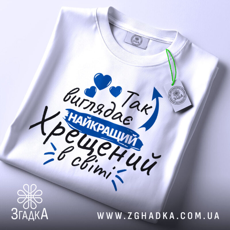 Футболка найкращий хрещений, біла, зображена зверху, фон світло-сірий, принт з текстом синього кольору, на футболці є етикетка.