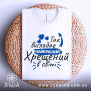 Футболка найкращий хрещений, біла, зображена на плетеній основі, фон білий, принт з текстом синього кольору та сердечками.