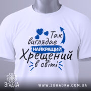 Футболка найкращий хрещений, біла, зображена зверху, фон світло-сірий, принт з текстом синього кольору, на футболці є етикетка.