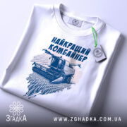 Футболка найкращий комбайнер, білого кольору, зображена в складеному вигляді на поверхні, фон світло-сірий, принт з комбайном та написом виконаний у темно-синьому кольорі, на футболці є етикетка з зеленим шнурком.