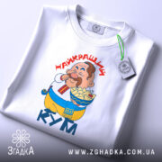 Футболка найкращий кум, біла, складена на поверхні, фон світло-сірий, принт з персонажем, що їсть, текст "НАЙКРАЩИЙ КУМ" червоним шрифтом, на ярлику зелена стрічка.