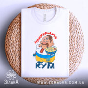 Футболка найкращий кум, біла, складена на плетеній кошику, фон світло-сірий, принт з персонажем, що їсть, текст "НАЙКРАЩИЙ КУМ" червоним шрифтом, видно етикетку на комірці.