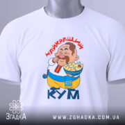 Футболка найкращий кум, біла, складена на поверхні, фон світло-сірий, принт з персонажем, що їсть, текст "НАЙКРАЩИЙ КУМ" червоним шрифтом, на ярлику зелена стрічка.