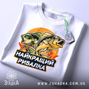 Футболка найкращий рибалка, біла, лежить на поверхні, злегка згорнута, фон світло-сірий, принт з рибами та написом, на футболці є етикетка з зеленим шнурком.