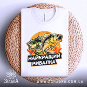 Футболка найкращий рибалка, біла, складена на плетеній основі, фон світло-сірий, принт з рибами та написом, видно етикетку на горловині.