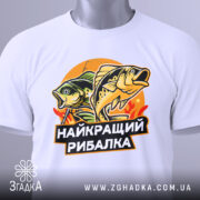 Футболка найкращий рибалка, біла, лежить на поверхні, злегка згорнута, фон світло-сірий, принт з рибами та написом, на футболці є етикетка з зеленим шнурком.