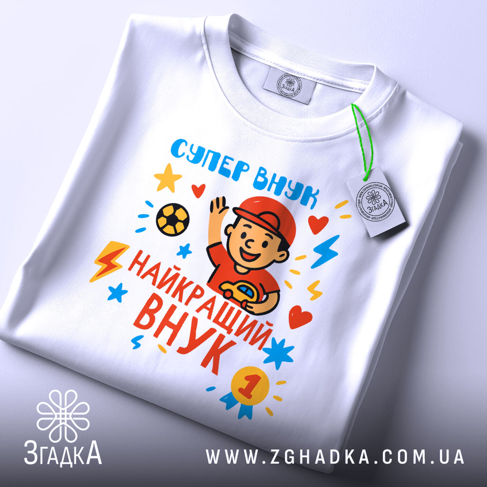 Футболка найкращий супер внук, біла, складена на столі, фон світло-сірий, принт з веселим хлопчиком у червоній кепці, який махає рукою, яскраві кольори принта: червоний, жовтий, блакитний, з зірочками і блискавками, на футболці є етикетка з зеленим шнурком.