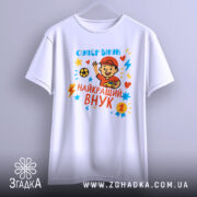 Футболка найкращий супер внук, біла, зображена на манекені з середнього ракурсу, фон світло-сірий, принт з веселим хлопчиком у червоній кепці, який тримає м'яч і машинку, яскраві кольори принта: червоний, жовтий, блакитний, з зірочками і блискавками.