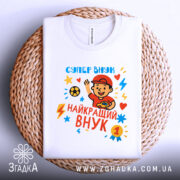 Футболка найкращий супер внук, біла, складена на плетеній основі, фон світло-сірий, принт з веселим хлопчиком у червоній кепці, який махає рукою, яскраві кольори принта: червоний, жовтий, блакитний, з зірочками і блискавками.