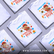 Футболка найкращий супер внук, біла, зображена на манекені з ближнього ракурсу, фон світло-сірий, принт з веселим хлопчиком у червоній кепці, який тримає м'яч і машинку, яскраві кольори принта: червоний, жовтий, блакитний, з зірочками і блискавками.