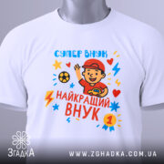 Футболка найкращий супер внук, біла, складена на столі, фон світло-сірий, принт з веселим хлопчиком у червоній кепці, який махає рукою, яскраві кольори принта: червоний, жовтий, блакитний, з зірочками і блискавками, на футболці є етикетка з зеленим шнурком.