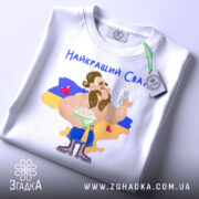 Футболка найкращий сват, білого кольору, складена на столі, фон світлий, принт з мультяшним персонажем, що тримає тарілку з варениками, на фоні жовто-блакитний прапор України, текст "Найкращий Сват" зверху, на футболці є етикетка.