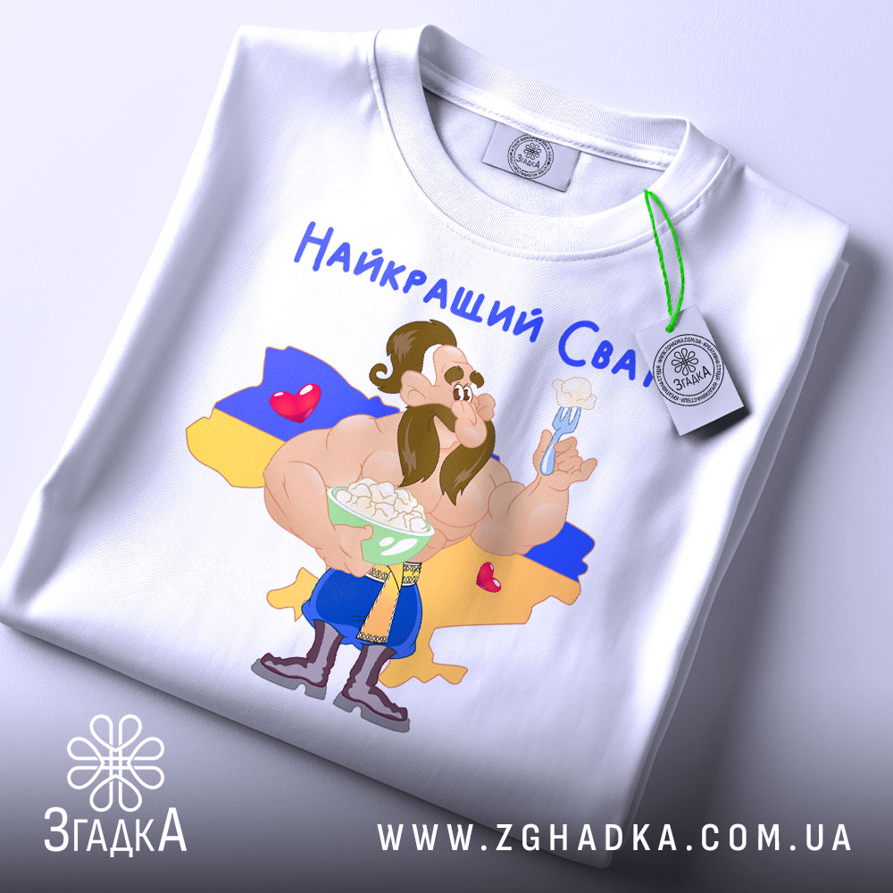 Футболка найкращий сват, білого кольору, складена на столі, фон світлий, принт з мультяшним персонажем, що тримає тарілку з варениками, на фоні жовто-блакитний прапор України, текст "Найкращий Сват" зверху, на футболці є етикетка.