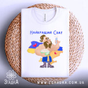 Футболка найкращий сват, білого кольору, складена на плетеній основі, фон нейтральний, принт з мультяшним персонажем, що тримає тарілку з варениками, на фоні жовто-блакитний прапор України, текст "Найкращий Сват" зверху.