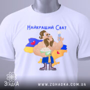 Футболка найкращий сват, білого кольору, складена на столі, фон світлий, принт з мультяшним персонажем, що тримає тарілку з варениками, на фоні жовто-блакитний прапор України, текст "Найкращий Сват" зверху, на футболці є етикетка.