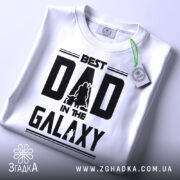 Футболка найкращий тато, біла, складена на поверхні, фон світло-сірий, принт чорний з написом 'BEST DAD IN THE GALAXY' та силуетами персонажів, з етикеткою на зеленому шнурку.