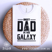 Футболка найкращий тато, біла, складена на круглій кошику, фон світло-сірий, принт чорний з написом 'BEST DAD IN THE GALAXY' та силуетами персонажів.
