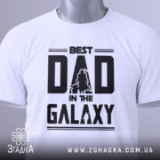 Футболка найкращий тато, біла, складена на поверхні, фон світло-сірий, принт чорний з написом 'BEST DAD IN THE GALAXY' та силуетами персонажів, з етикеткою на зеленому шнурку.