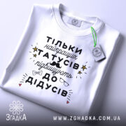 Футболка найкращий татусь, білого кольору, складена на столі, фон світло-сірий, принт чорного кольору з жовтими зірочками та малюнками окулярів і вусів, на комірці є етикетка.