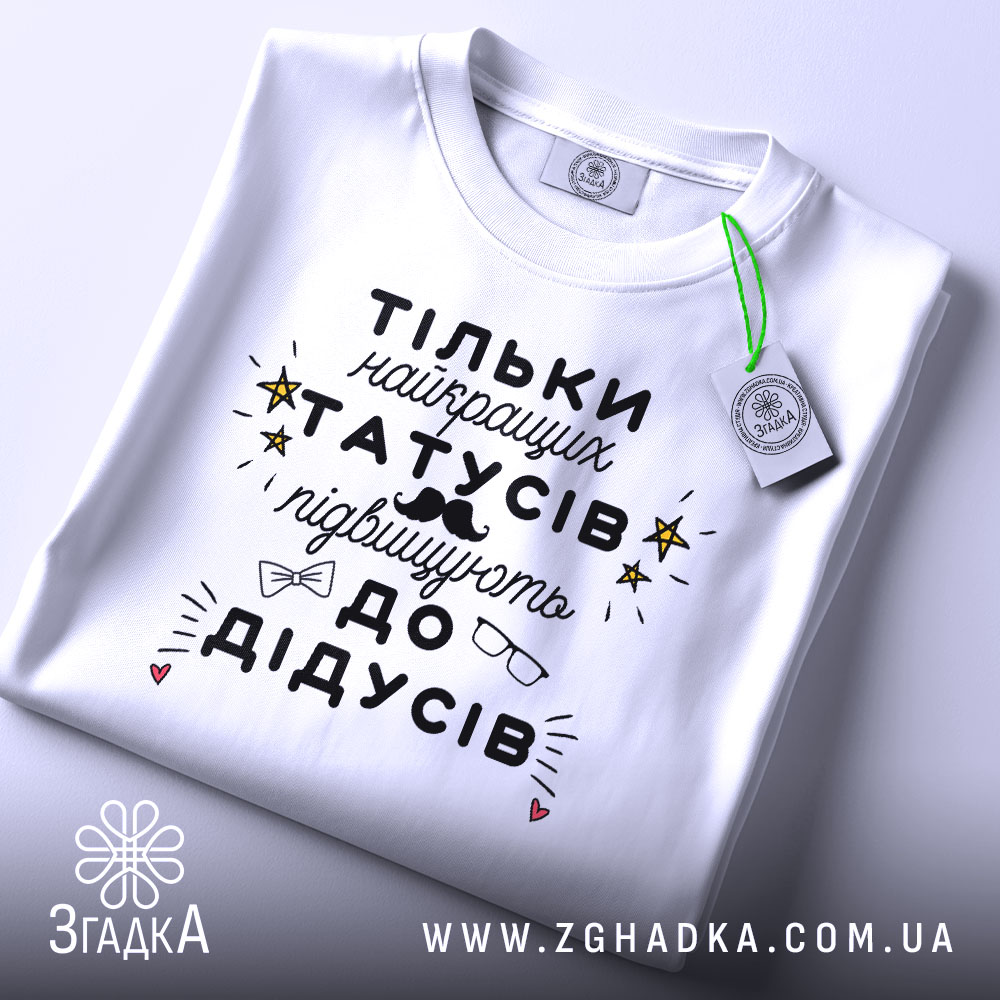 Футболка найкращий татусь, білого кольору, складена на столі, фон світло-сірий, принт чорного кольору з жовтими зірочками та малюнками окулярів і вусів, на комірці є етикетка.