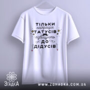 Футболка найкращий татусь, білого кольору, підвішена на вішалці, фон світло-сірий, принт чорного кольору з жовтими зірочками та малюнками окулярів і вусів.
