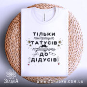 Футболка найкращий татусь, білого кольору, складена на плетеній кошику, фон світло-сірий, принт чорного кольору з жовтими зірочками та малюнками окулярів і вусів.