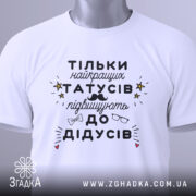 Футболка найкращий татусь, білого кольору, складена на столі, фон світло-сірий, принт чорного кольору з жовтими зірочками та малюнками окулярів і вусів, на комірці є етикетка.