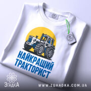 Футболка найкращий тракторист. Біла футболка з коротким рукавом, зображення трактора на жовтому фоні, текст 'НАЙКРАЩИЙ ТРАКТОРИСТ' синього кольору під принтом, футболка лежить на поверхні, видно етикетку з зеленим шнурком.