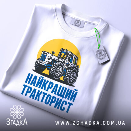 Футболка найкращий тракторист. Біла футболка з коротким рукавом, зображення трактора на жовтому фоні, текст 'НАЙКРАЩИЙ ТРАКТОРИСТ' синього кольору під принтом, футболка лежить на поверхні, видно етикетку з зеленим шнурком.