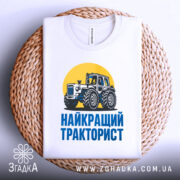 Футболка найкращий тракторист. Біла футболка з коротким рукавом, зображення трактора на жовтому фоні, текст 'НАЙКРАЩИЙ ТРАКТОРИСТ' синього кольору під принтом, футболка лежить на плетеній основі, фон світлий.