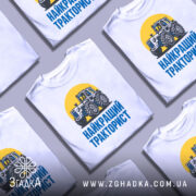 Футболка найкращий тракторист. Біла футболка з коротким рукавом, зображення трактора на жовтому фоні, текст 'НАЙКРАЩИЙ ТРАКТОРИСТ' синього кольору під принтом, фон нейтральний, футболка представлена на манекені.