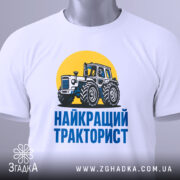 Футболка найкращий тракторист. Біла футболка з коротким рукавом, зображення трактора на жовтому фоні, текст 'НАЙКРАЩИЙ ТРАКТОРИСТ' синього кольору під принтом, футболка лежить на поверхні, видно етикетку з зеленим шнурком.